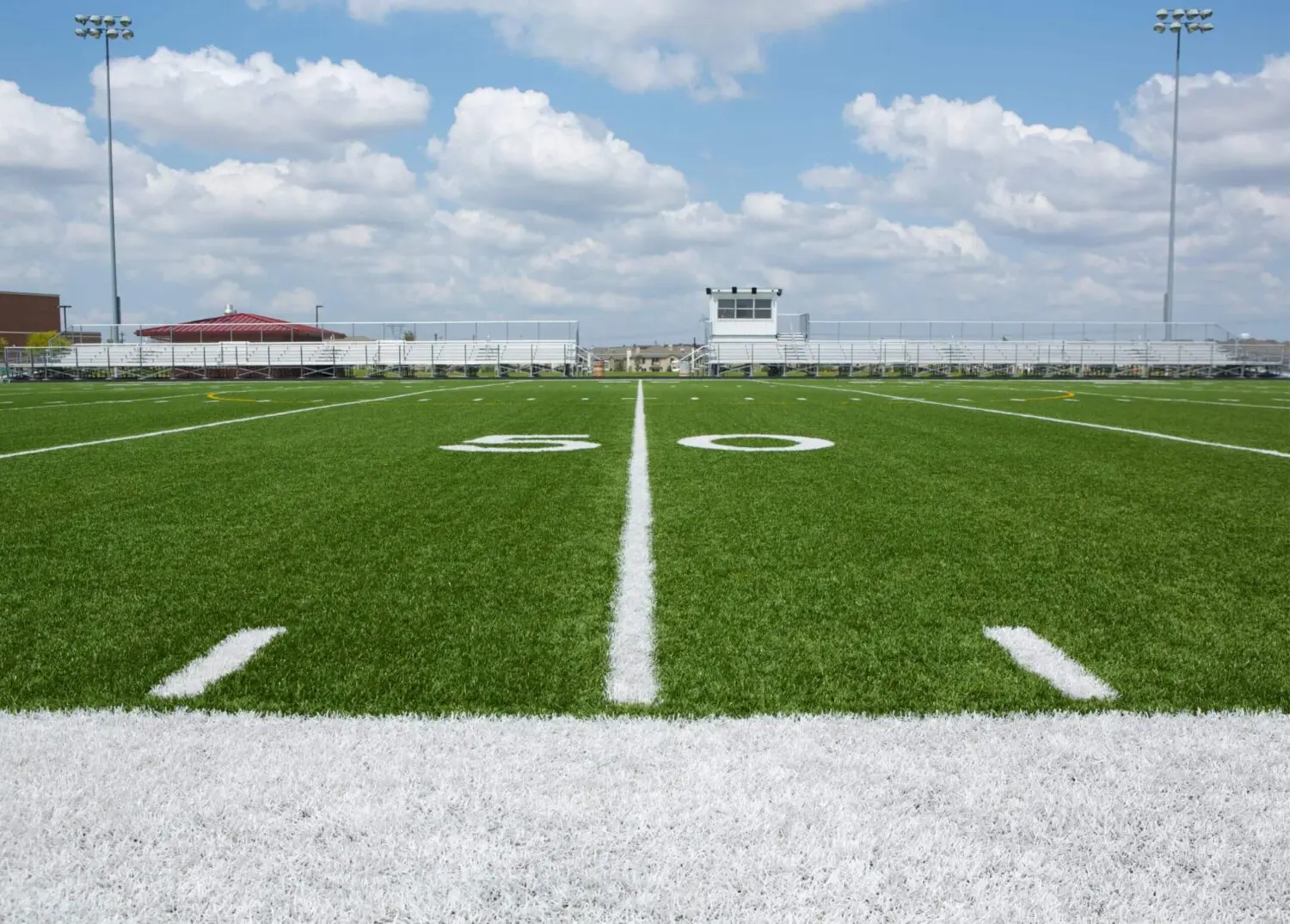 A football field stretches before you, its Vegas Artificial Turf radiating a vibrant green. The crisp white 50-yard line marks the center, while bleachers and a small building stand under a partly cloudy sky in the background.