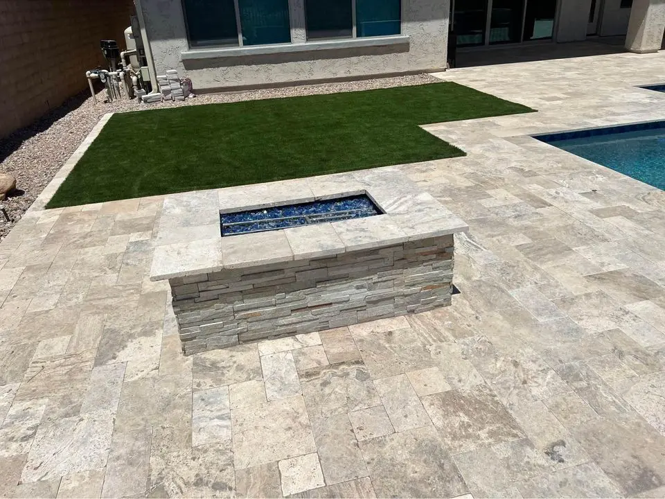 Stone patio area featuring a rectangular fire pit with blue glass stones. Adjacent are a patch of green artificial grass and a swimming pool. The backdrop is a light-colored building with large windows.