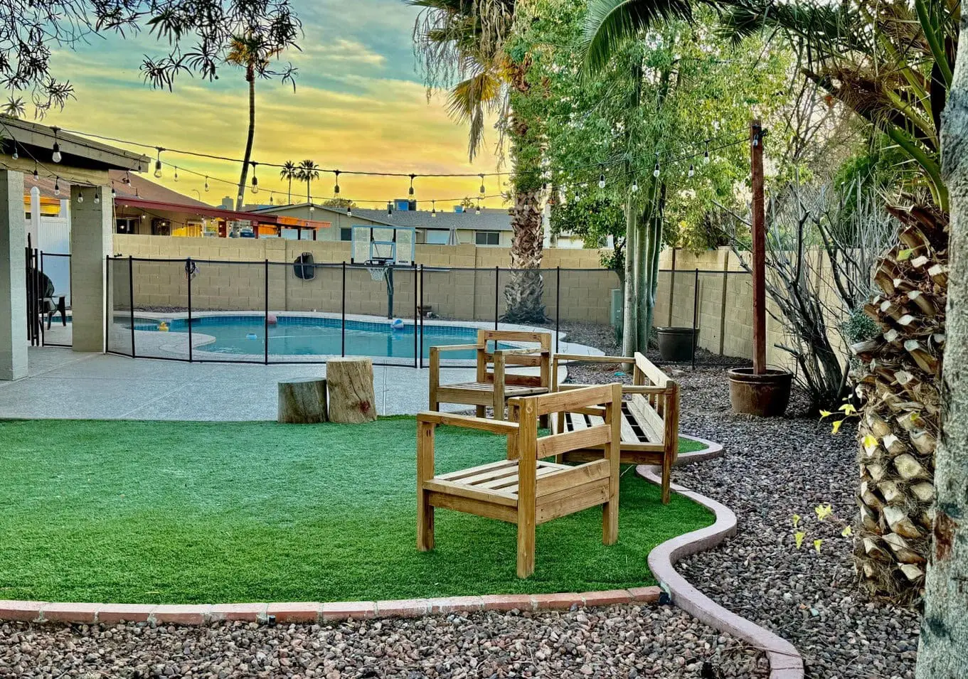 The backyard features two wooden benches on a cozy Vegas Artificial Turf patch, enclosed by gravel. A fenced-in pool shimmers nearby, with string lights twinkling above and lush greenery all around. The evening sky is painted in shades of blue and yellow, hinting at sunset.