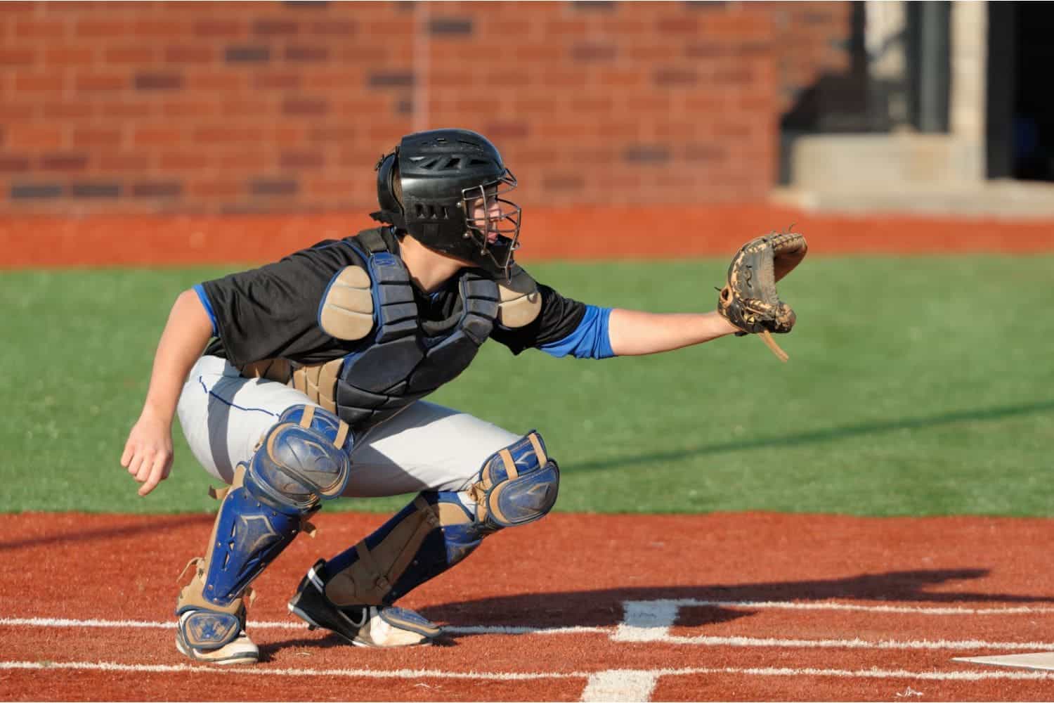 A baseball catcher in protective gear crouches on the lush green field, extending their glove forward. The player wears a black helmet, chest protector, and blue shin guards, with the vibrant Vegas artificial turf and brick wall creating a striking backdrop.